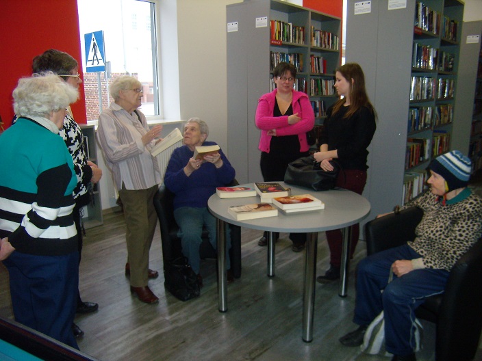 Wnętrze biblioteki, przy stoliku siedzą panie z Klubu Seniora zwiedzają oddział dla dorosłych.