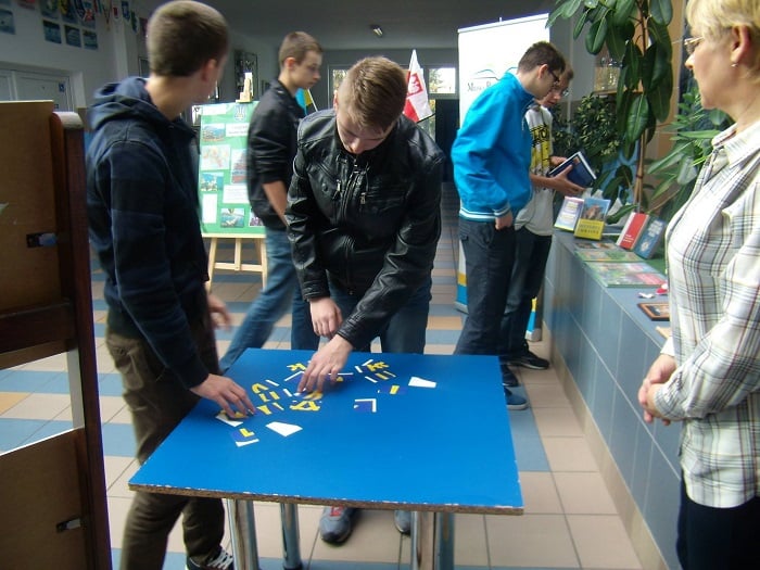 Wnętrze biblioteki w Szczytnie przy stoliku stoi młodzież rozwiązuje zadanie z gry miejskiej.