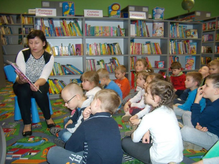 Przy regale z książkami siedzi grupa dzieci , słuchają bajki , którą czyta bibliotekarka Ania.