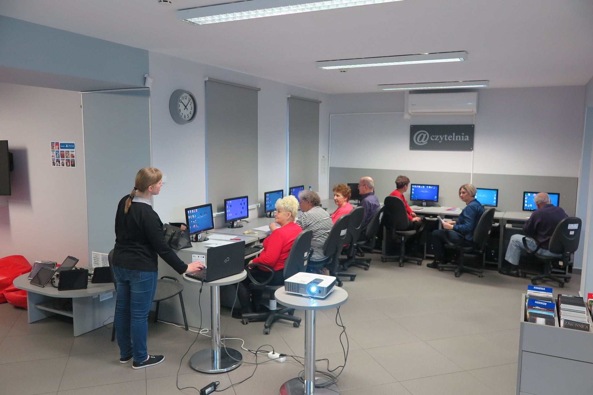 Wnętrze biblioteki (miediateka) bibliotekarka Małgorzata Trąmpczyńska prowadzi kurs komputerowy. Siedmioro śmiałków siedzi przed komputerami na kolejnej edycji kursu komputerewego z podstawowej obsługi komputera i usług internetowych. 