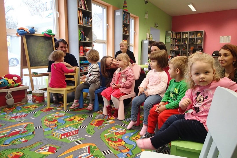 Wnętrze biblioteki. Na krzesełkach siedzi grupa dzieci z opiekunami.