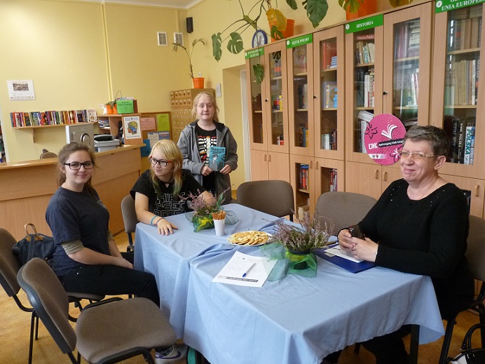 Wnętrze biblioteki, przy stoliku siedzi pani Ewa Sotomska i trzy członkinie DKK wspólnie omawiają książkę Grzegorza Gortata „Miasteczko ostatnich westchnień”.