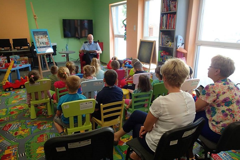 Grupa dzieci siedzi na krzesełkach w bibliotece uważnie słuchają jak czyta strażak Państwowej Straży Pożarnej w Działdowie - Leszek Góralski książkę "Jak Wojtek został strażakiem" z okazji  XIV Ogólnopolskiego Tygodnia Czytania Dzieciom.