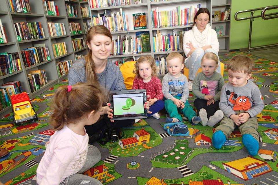 Na kolorowej wykładzinie siedzą dzieci biorą udział w lekcji bibliotecznej, Bibliotekarka Aleksandra pokazuje na tablecie owoce kiwi.