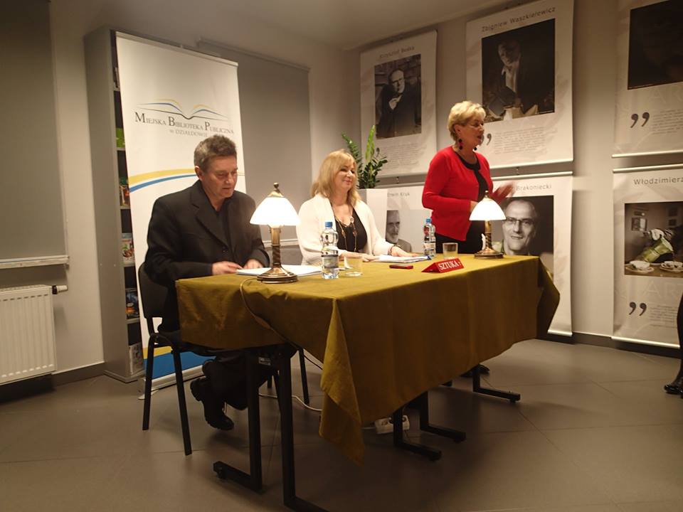 Wnętrze biblioteki, przy stoliku siedzą aktorzy  Teatru Stefana Jaracza w Olsztynie Alicja Kochańska i Artur Steranko obok pani Elżbieta Lenkiewicz, za nimi baner MBP.