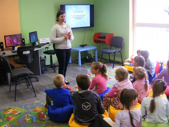 Grupa dzieci siedzi na kolorowych pufach, bibliotekarka Ania prowadzi lekcję na temat Unii Europejskiej w tle telewizor z prezentacją.