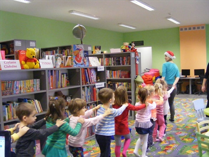 Wnętrze biblioteki. Grupa dzieci tańczy w utworzonym pociągu z bibliotekarką Aleksandrą Cybulską obok regały z książkami.
