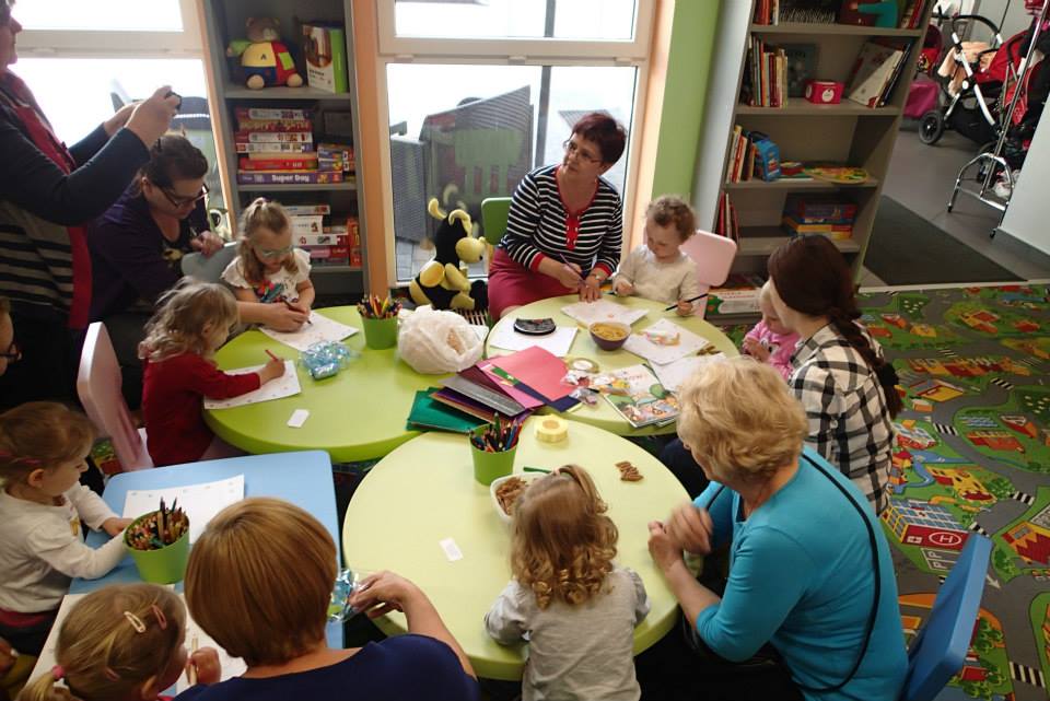 Przy stolikach siedzą dzieci z opiekunkami wspólnie wykonują prace plastyczne.