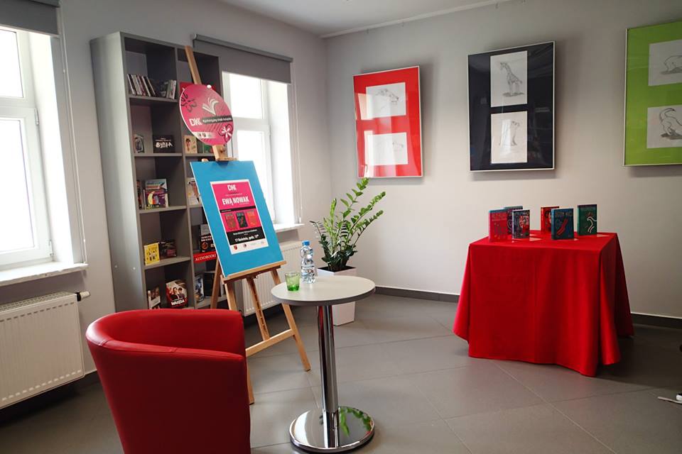 Stolik z książkami autorki Ewy Nowak , czerwony fotel, stolik z butelką wody i szklanką oraz sztaluga z plakatem spotkania z autorką Ewą Nowak.