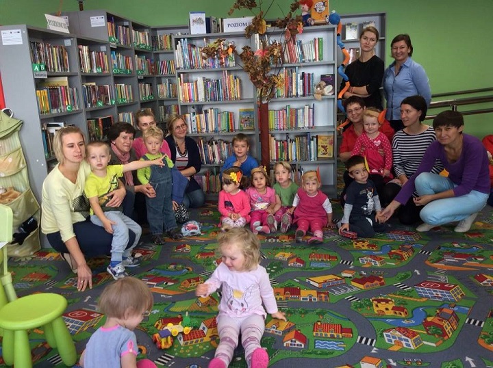 Przy regałach z książkami siedzą dzieci z opiekunami świętują pierwszy dzień jesieni. 