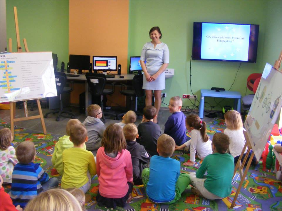 Wnętrze biblioteki, grupa dzieci siedzi na kolorowej wykładzinie i bierze udział w zajęciach o unii europejskiej, na sztaludze znajduje się krzyżówka o unii a na telewizorze prezentacja o unii europejskiej.