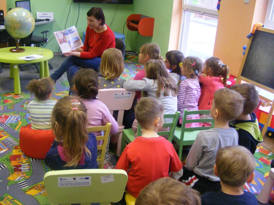 Na krzesełkach siedzą dzieci, bibliotekarka Ania czyta im książeczke, w tle na stoliku stoi globus.