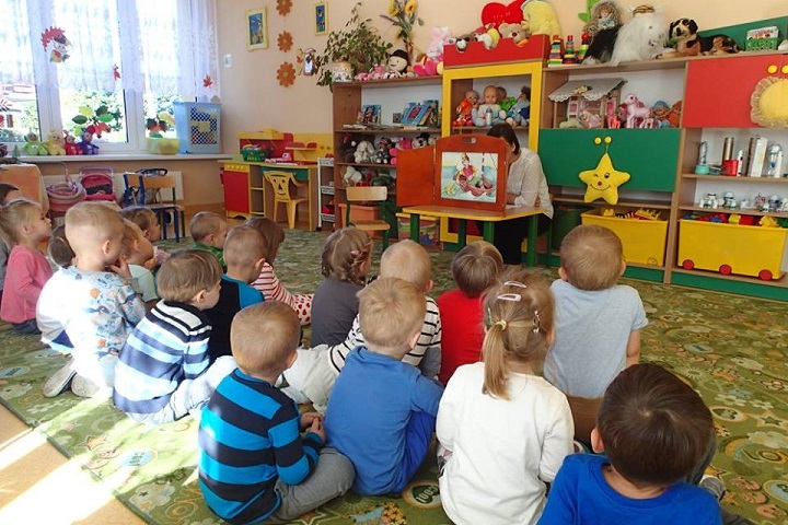 Wnętrze Przedszkola nr 3 na kolorowej wykładzinie siedzą dzieci, bibliotekarka Ania Wiśniewska czyta teatrzyk kamishibai "O rybaku i złotej rybce".