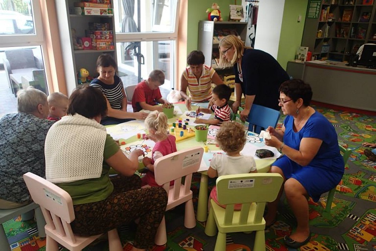 Przy stoliku siedzą dzieci z opiekunami wspólnie malują farbami.