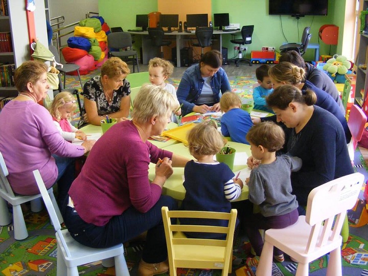Przy stolikach siedzą dzieci z opiekunkami wspólnie wykonują prace plastyczne.