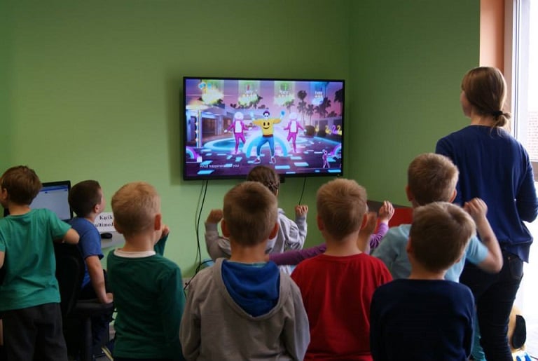 Grupa dzieci z opiekunką tańczą do wybranej muzyki, w tle telewizor z prezentacją zabawy.