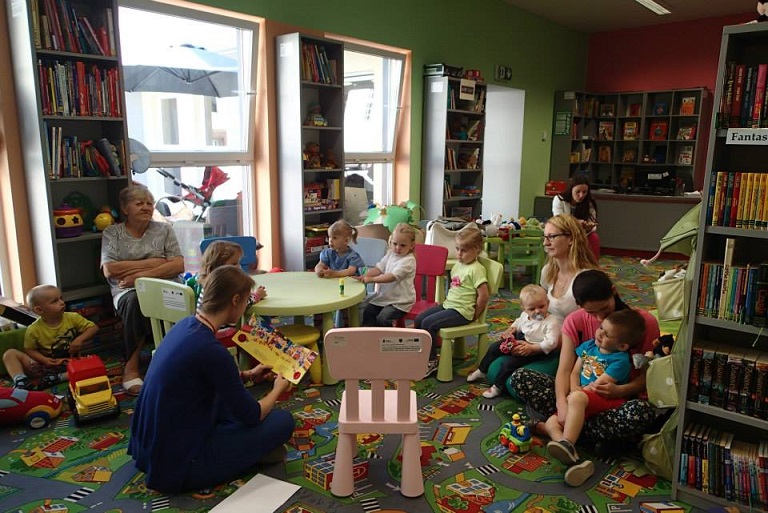 Grupa dzieci siedzi przy stoliku z opiekunami, bibliotekarka Małgorzata Trąmpczyńska czyta  wiersze Juliana Tuwima „Lokomotywa” i „Rzepka”. 