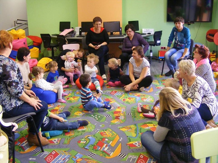 Na kolorowej wykładzinie siedzą dzieci z opiekunkami, bibliotekarka Ania prowadzi zajęcia. 