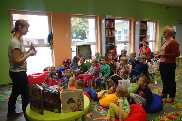Na krzesełkach siedzą dzieci, bibliotekarka Aleksandra opowiada   o drodze książki od pisarza do czytelnika. Na stoliku stoją wybrane książki dla dzieci. 