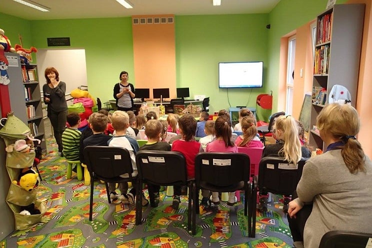 Na krzesełkach siedzą uczniowie z ZS w Księżym Dworze biorą udział w pierwszej lekcji bibliotecznej na temat powstawania książek, zajęcia prowadzi bibliotekarka Ania w tle telewizor z prezentacją. 
