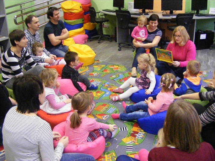 Na kolorowej wykładzinie siedzą dzieci z opiekunami wspólnie słuchają opowiadania  pt. " Skąd się biorą prezenty". 