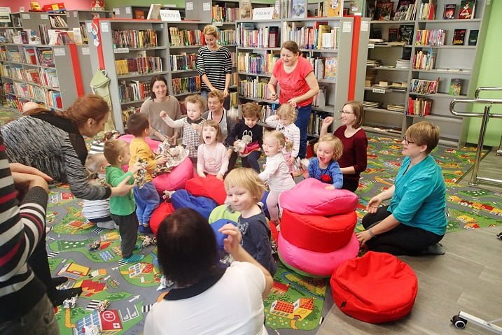 Grupa dzieci z opiekunami podczas zajęć w Klubie Malucha,w tle regały z książkami.