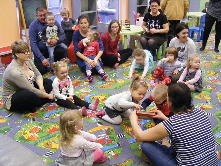 Wnętrze biblioteki. Na kolorowej wykładzinie siedzą dzieci z opiekunami, bibliotekarka Ania pokazuje książkę o Świętym Mikołaju.