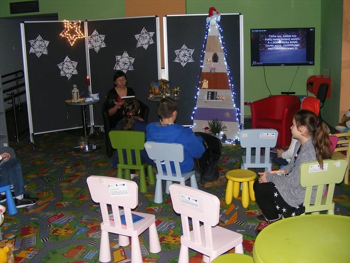 Na krzesełkach siedzą dzieci, bibliotekarka Ania Wiśniewska czyta  "Opowieść wigilijną" w tle stoi choinka, prezentacja świąteczna na telewizorze oraz białe gwiazdki przyczepione na tablicach.