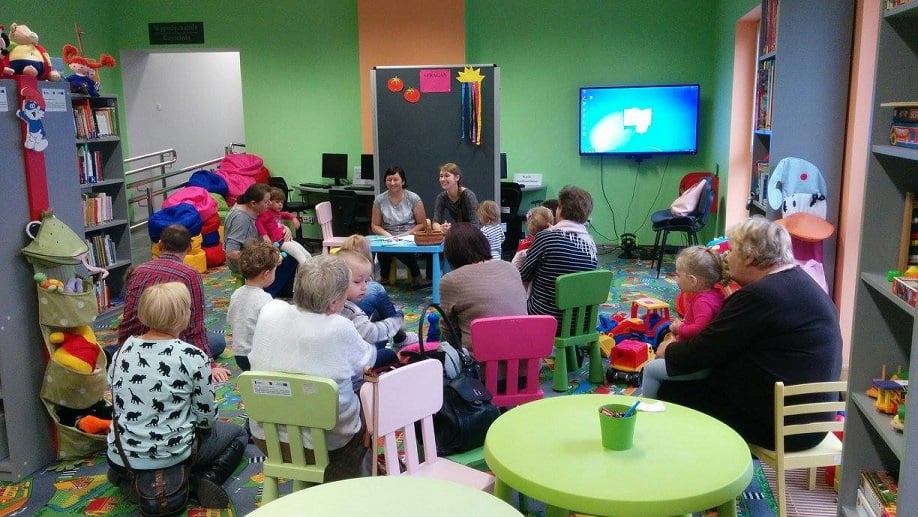 Na krzesełkach siedzą dzieci z opiekunami wspólnie biorą udział w zabawie z bibliotekarkami Anią i Aleksandrą, razem poznają warzywa.