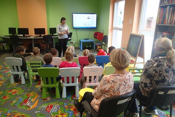 Na krzesełkach siedzą dzieci z opiekunkami, bibliotekarka Ania opowiada jak powstaje książka, w tle na telewizorze wyświetla się prezentacja o książce.