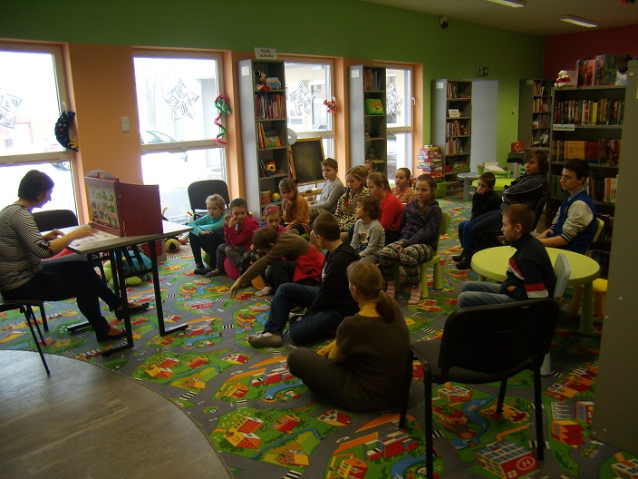 Wnętrze biblioteki. Grupa dzieci słucha teatrzyku Kamishibai, czyta bibliotekarka Ania.