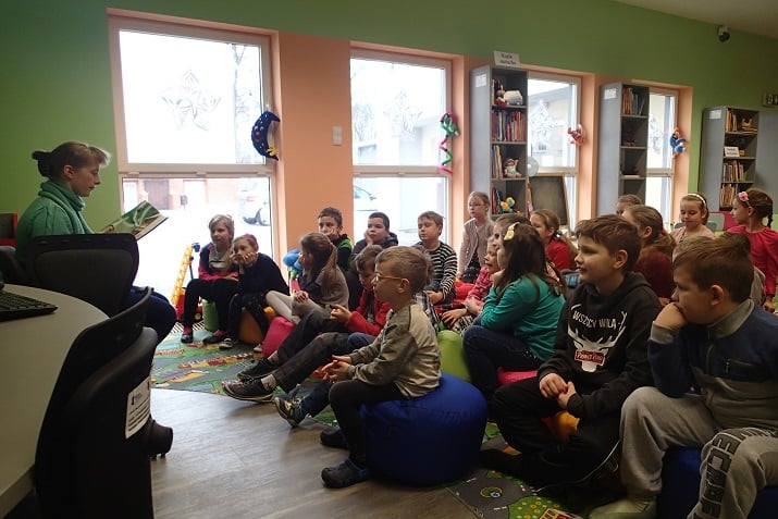 Grupa dzieci siedzi na pufach, bibliotekarka Małgorzata czyta dzieciom książkę.