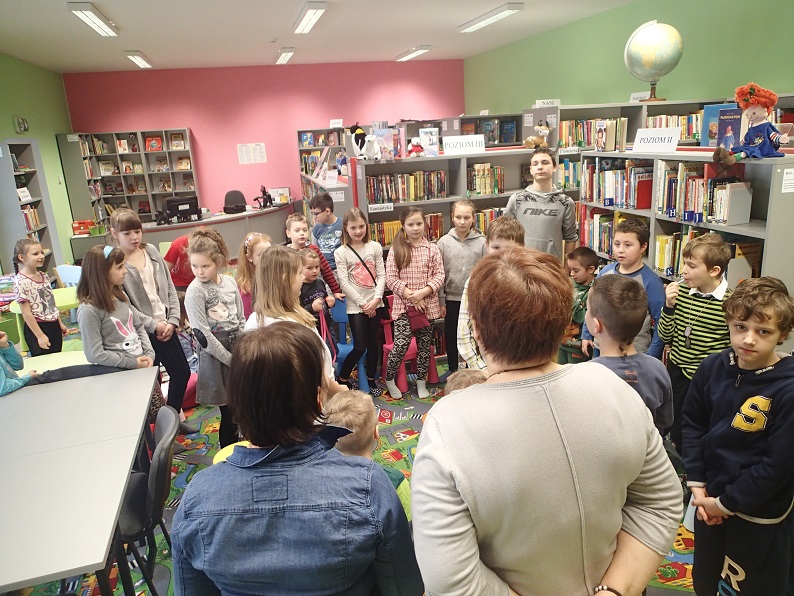 Grupa dzieci z bibliotekarką wspólnie biorą udział w zabawie, w tle regały z książkami.