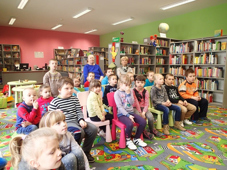 Wnętrze biblioteki, grupa dzieci siedzi na krzesełkach z opiekunami w tle regały z książkami.
