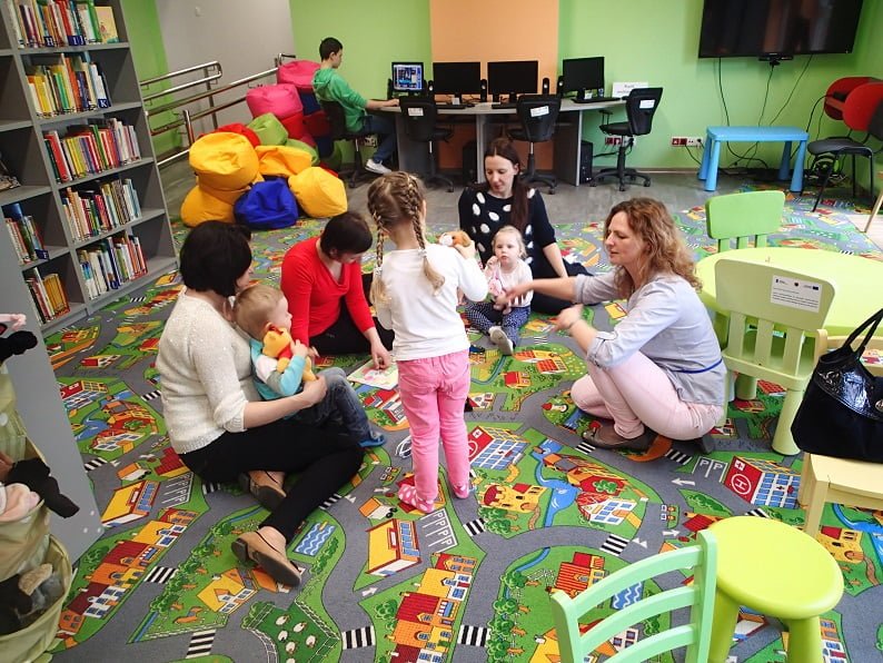 Na kolorowej wykładzinie siedzą dzieci z opiekunkami, bibliotekarka Ania czyta  dzieciom bajki J.Tuwima "Okulary" i "Zosia samosia". 