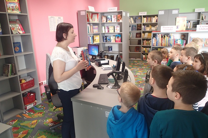 Bibliotekarka Ania pokazuje książki dla dzieci przedszkolakom, w tle regały z książkami.