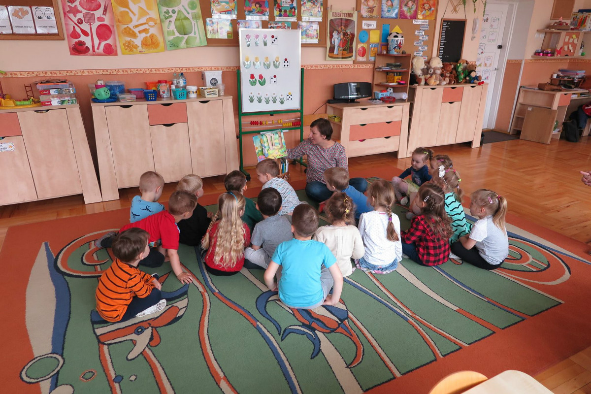 Wnętrze Przedszkola nr 1. Dzieci siedzą na kolorowym dywanie i słuchają opowiadania  "Złotowłosa" czyta bibliotekarka Ania Wiśniewska.