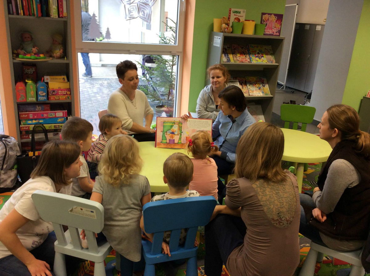 Wnętrze biblioteki. Przy stoliku siedzą dzieci z opiekunami, bibliotekarka Ania Wiśniewska czyta opowiadanie "Boże Narodzenie Tupcia Chrupcia". 