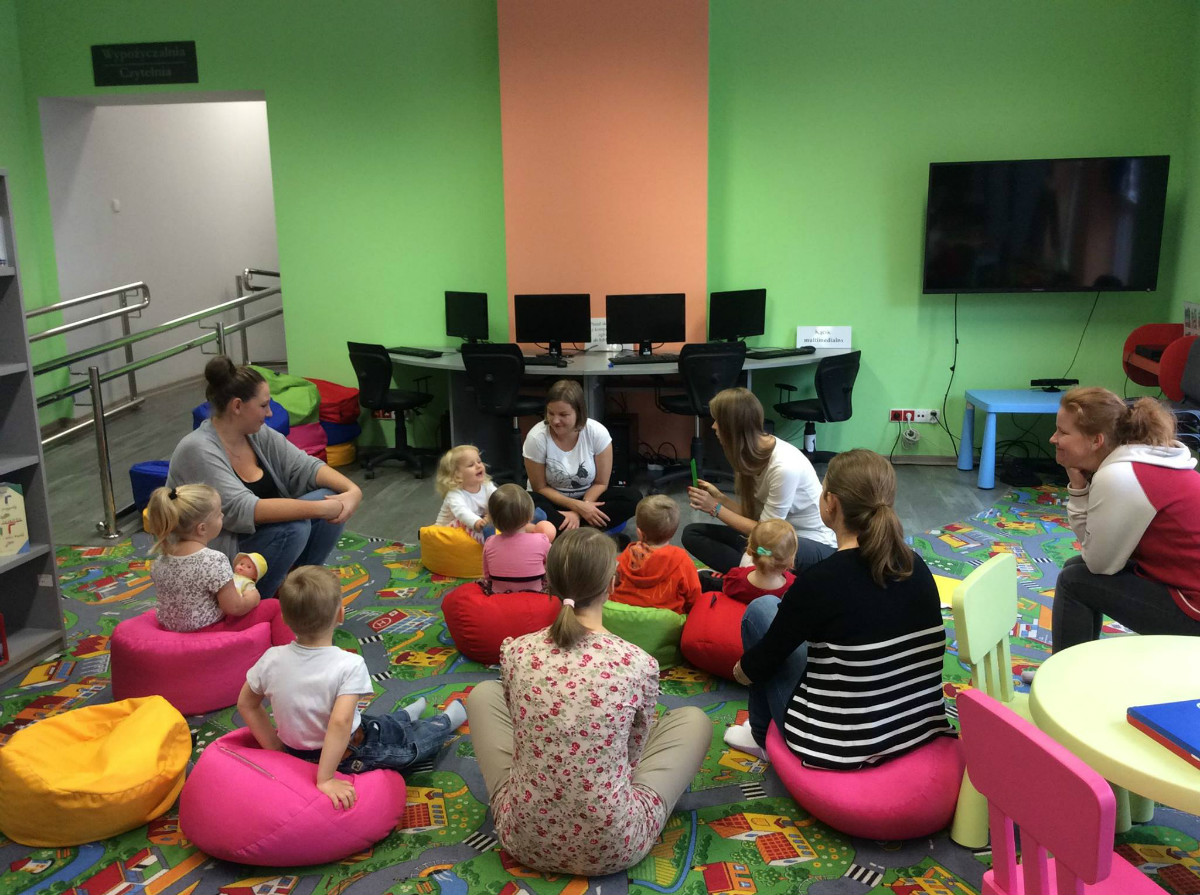 Wnętrze biblioteki. Na pufach siedzą dzieci  z opiekunkami,bibliotekarka Aleksandra Cybulska prowadzi zajęcia. 