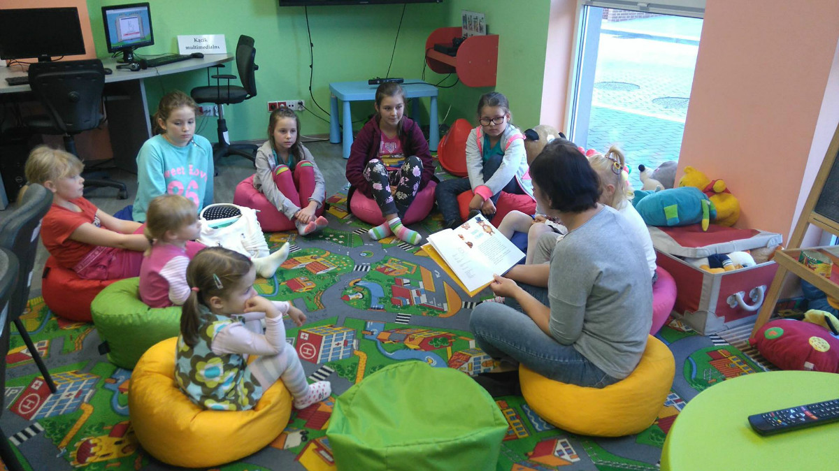 Wnętrze biblioteki. Na pufach siedzą dzieci, bibliotekarka Ania Wiśniewska czyta  opowiadania o Misiu Promyczku oraz Puchatku. 