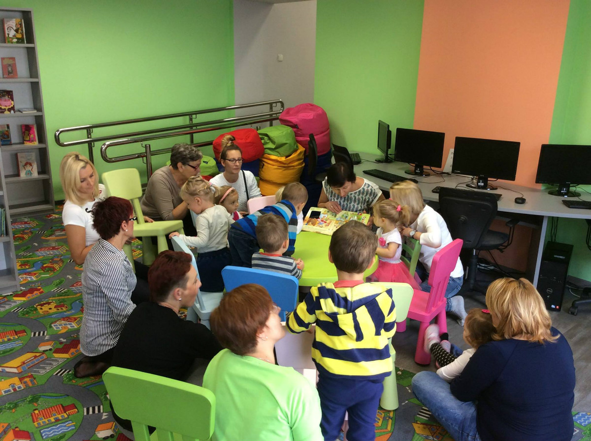 Wnętrze biblioteki. Przy stolikach siedzą dzieci z opiekunami, bibliotekarka Ania czyta opowiadanie "Kochane misie".