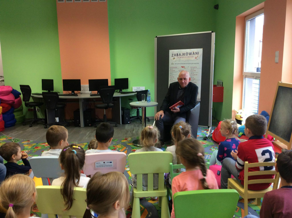 Wnętrze biblioteki. Na krzesełkach siedzą dzieci, burmistrz miasta Działdowo Grzegorz Mrowiński czyta bajkę „Kopciuszek”. 