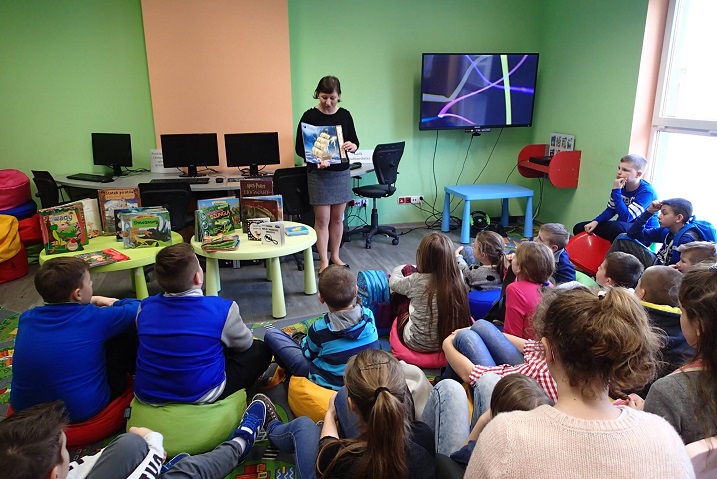 Na kolorowych pufach siedzą uczniowie ze SP w Białutach, , bibliotekarka Ania prowadzi lekcję na temat najciekawszych zasobów księgozbioru biblioteki.