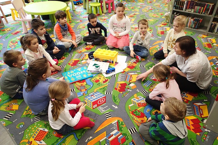 Na kolorowej wykładzinie w kółku siedzi grupa dzieci z bibliotekarką Anią Wiśniewską razem rozmawiają na temat  recyklingu odpadów z tworzyw sztucznych i gdzie wyrzucać takie odpady.