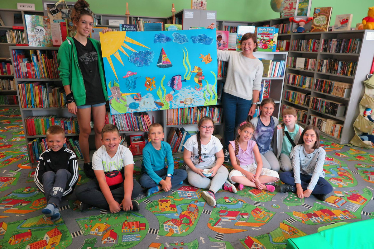 Wnętrze biblioteki, przy  regale z książkami morską pracę plastyczną dzieci trzyma w górze bibliotekarka Ania Wiśniewska z opiekunką, niżej na kolorowej wykładzinie siedzi ośmioro dzieci. 