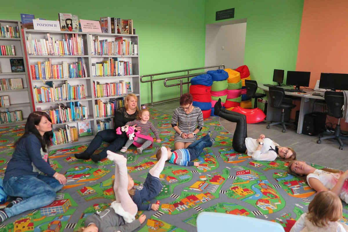 Wnętrze biblioteki. Grupa dzieci z opiekunami podczas zajęć w Klubie Malucha.