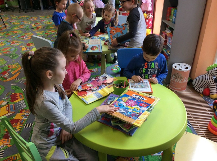 Przy zielonym stoliku siedzą dzieci oglądają książki z różnymi postaciami z bajek. 