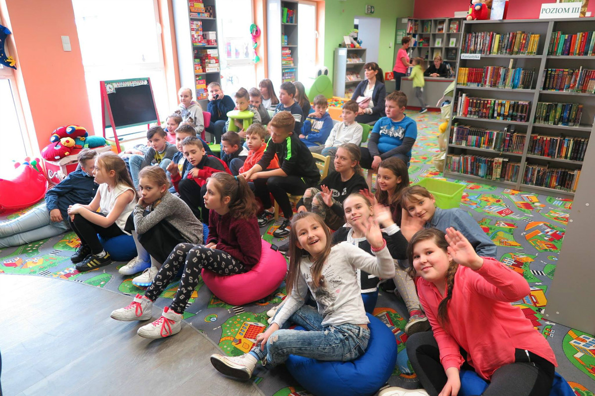 Wnętrze biblioteki. Na kolorowej wykładzinie siedzą dzieci z opiekunami, biorą udział w lekcji bibliotecznej. 