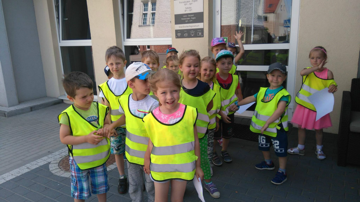 Na placu MBP stoi grupa dzieci z Przedszkola nr 3  w odblaskowych kamizelkach. 