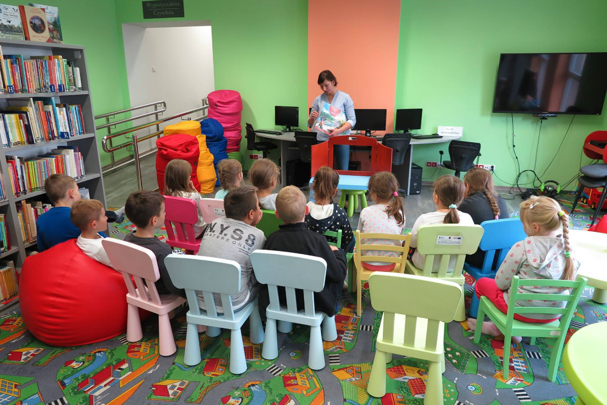 Wnętrze biblioteki, na krzesełkach siedzą dzieci słuchają teatrzyku Kamishibai "Kot w butach", zajęcia prowadzi bibliotekarka Ania Wiśniewska.